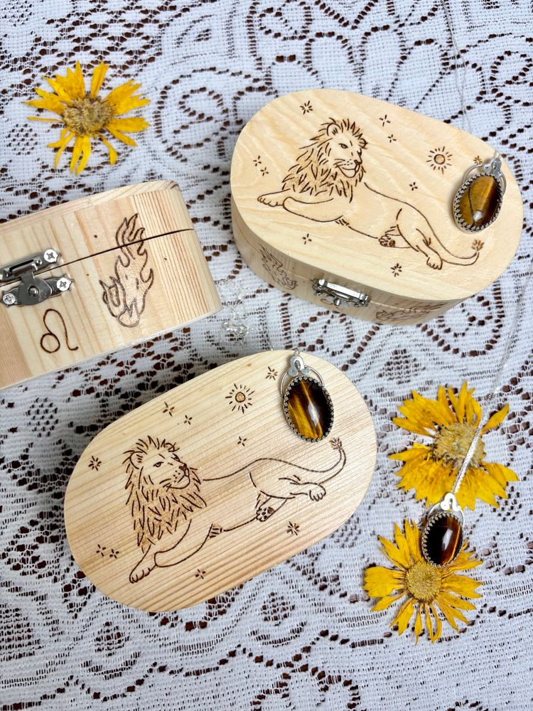 3 wood trinket boxes with pyrography of a lion, sparkles, a sun, flames, a Leo symbol and a Leo star constellation, 3 tiger’s eye silversmithed necklaces with a Leo symbol, displayed with dried dried flowers on a lace backdrop