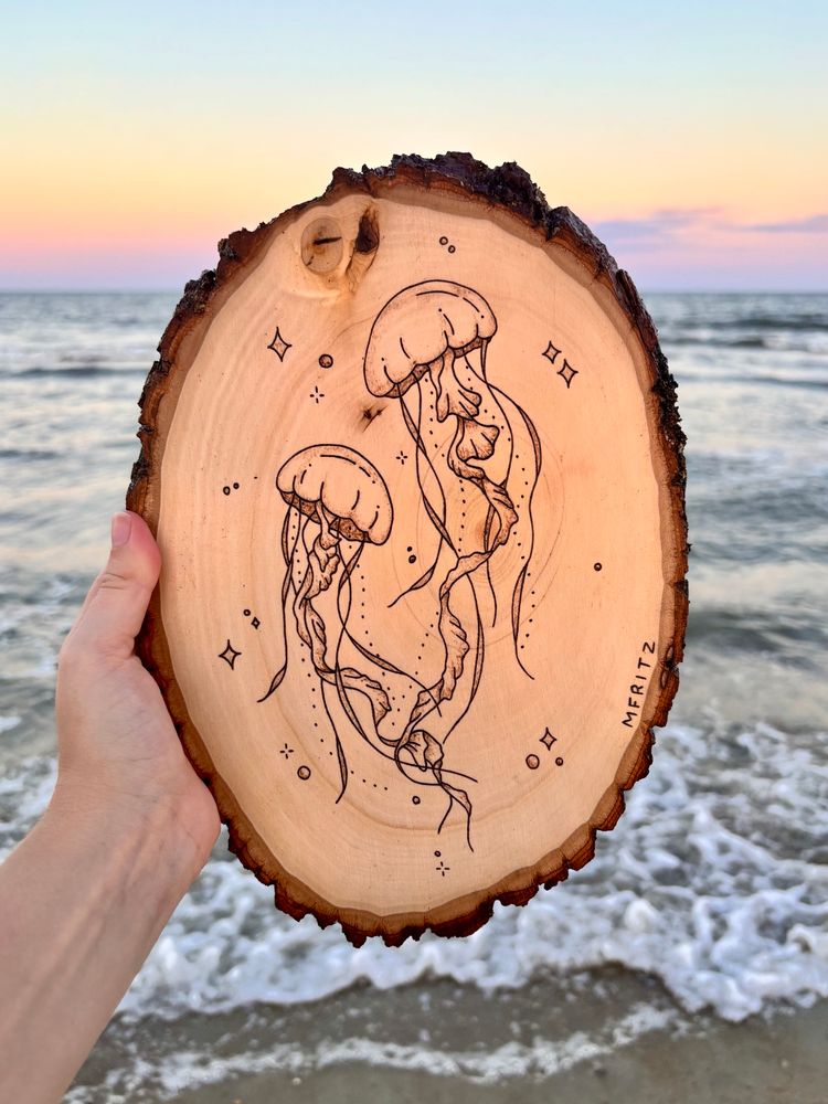 Pyrography of 2 jellyfish with bubbles and sparkles on an oval wood slice held in front of the Atlantic Ocean at sunset