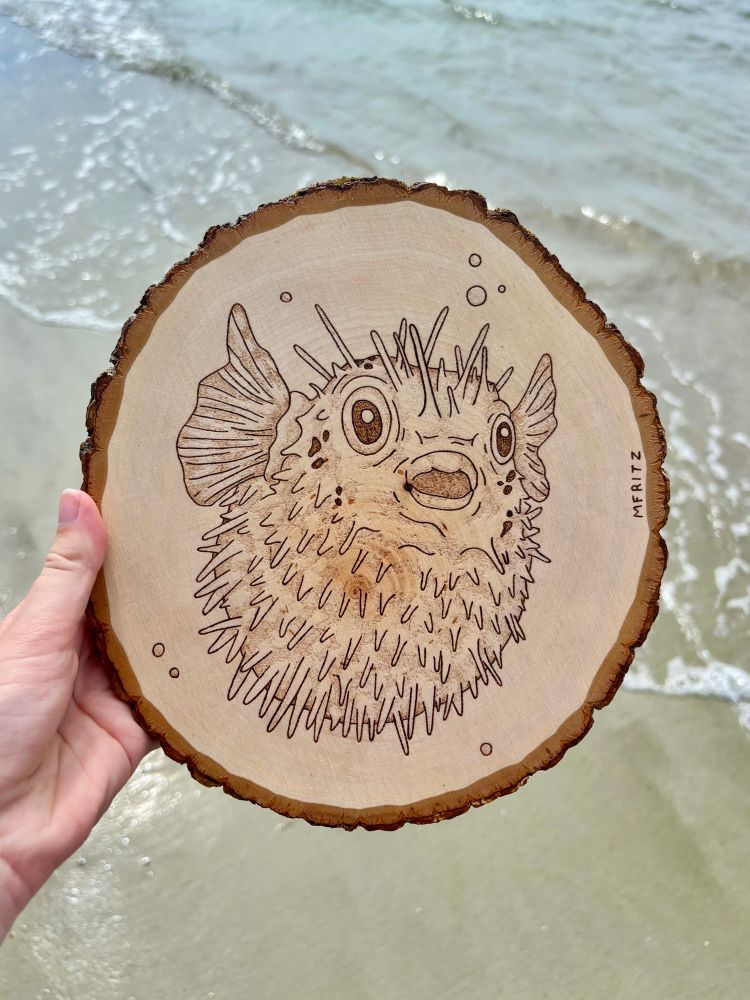 Pyrography of a pufferfish with bubbles on a round wood slice held in front of the sand and waves of the Atlantic Ocean