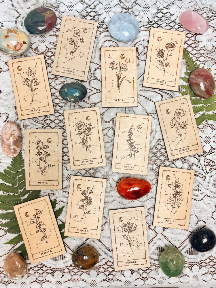 12 wood burned birth flower cards (snowdrop, violet, daffodil, daisy, lily of the valley, rose, larkspur, poppy, morning glory, marigold, chrysanthemum, holly), 12 crystal palmstones (polychrome jasper, sea jasper, blue calcite, rose quartz, labradorite, petrified wood, flower agate, carnelian, peach moonstone, golden healer, fluorite, black tourmaline) displayed on lace with dried ferns