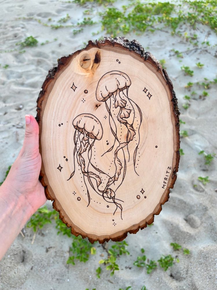 Pyrography of 2 jellyfish with sparkles and bubbles on an oval wood slice held in front of the sand with greenery
