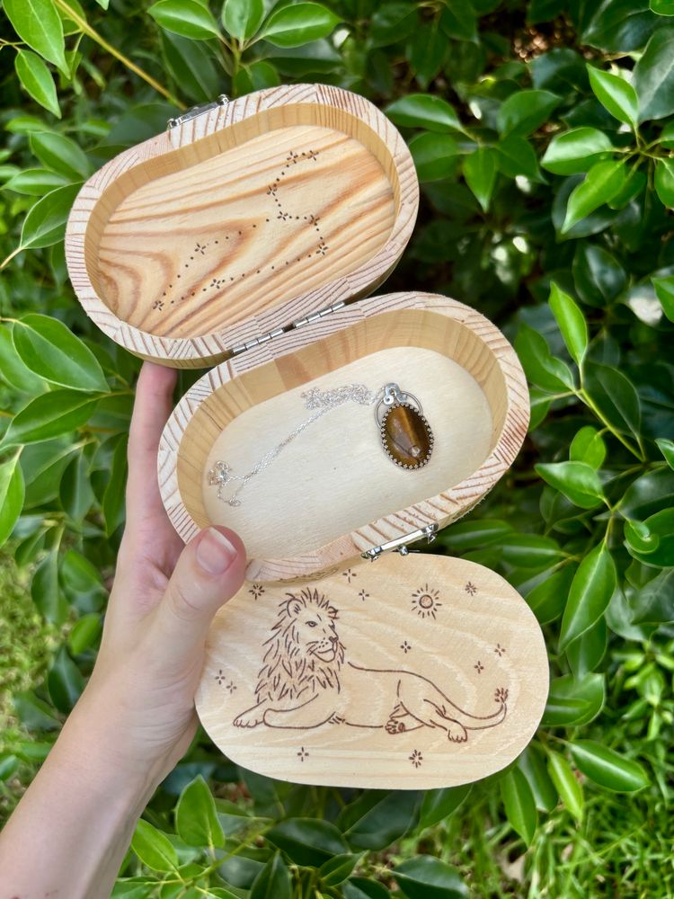 2 wood trinket boxes with pyrography of a lion, sparkles, a sun, flames, a Leo symbol and a Leo star constellation, 1 silversmithed Tiger’s Eye necklace with a Leo symbol held up in front of a leafy green backdrop