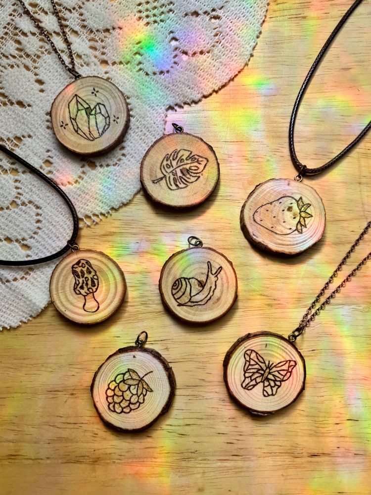 7 small wood slices with pyrography designs of crystals, monstera leaf, strawberry, morel mushroom, snail, raspberry and a butterfly with copper eye screws and jump rings attached for necklaces; faux leather waxed cord and copper chain. Displayed on a wood table with lace and some rainbow light reflections