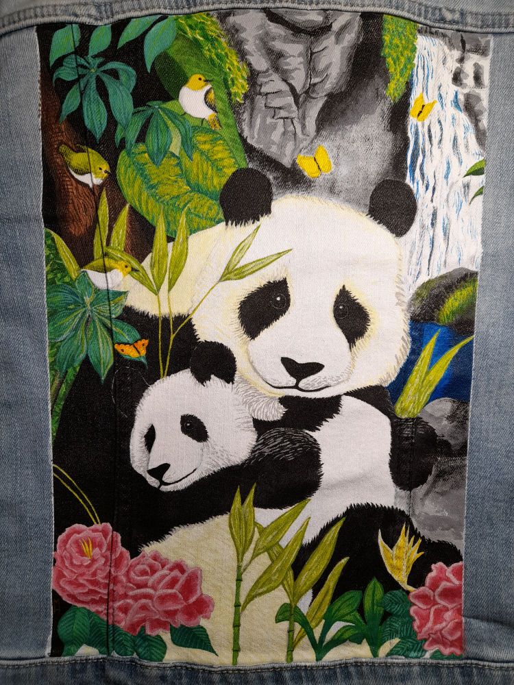 A painting on the back panel of a denim jacket. There are two panda bears, a mother and cub. The mother embraces the cub. They are surrounded by pink flowers, bright vegetation, birds, and butterflies. Behind them is a waterfall cascading down grey stone.