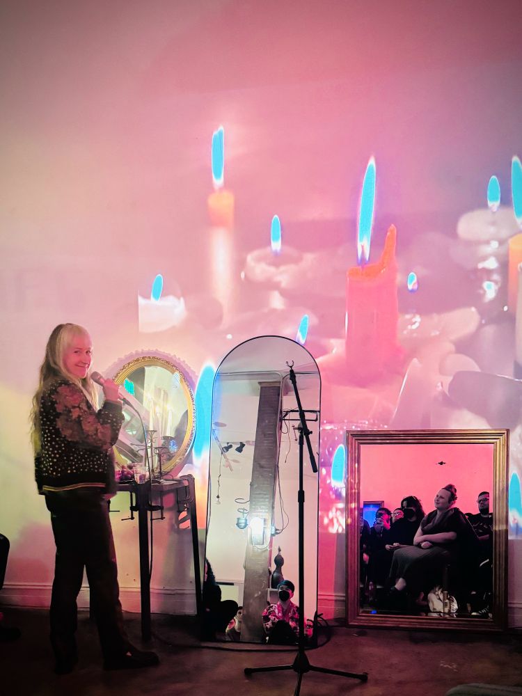 Poet holding microphone, standing in front of three mirrors and a wall of projected candles. 