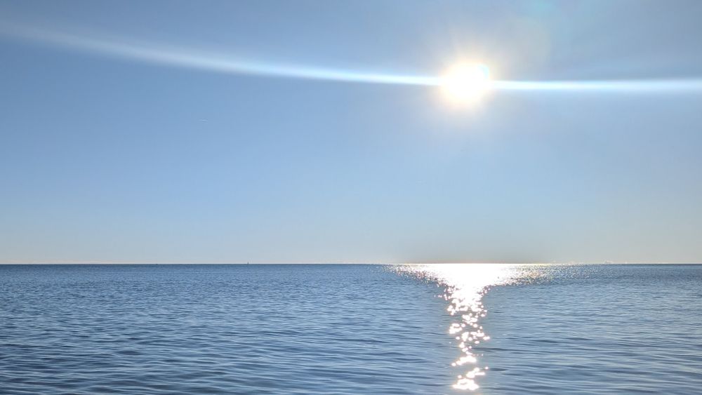 Bright sun reflecting on a calm sea with a pure horizon, under a clear blue sky.