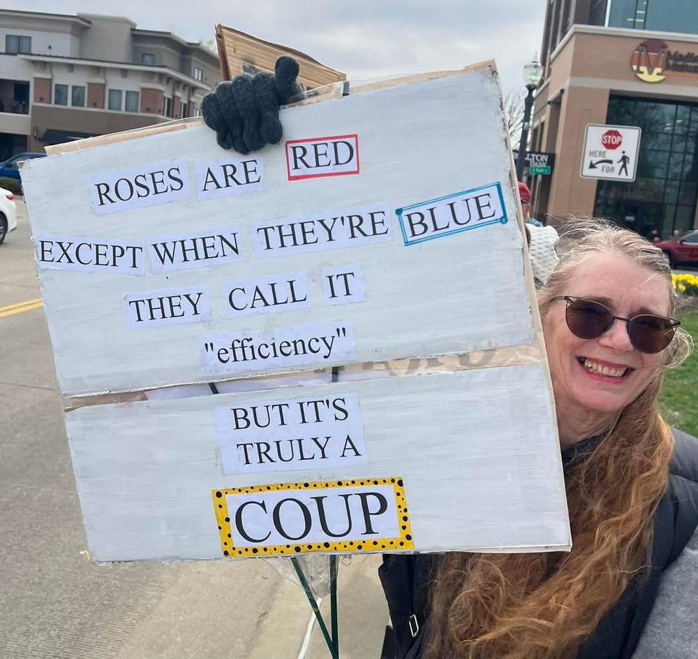 An old white lady (me) with long windblown greying ginger/auburn hair stands on the sidewalk next to a busy suburban street holding a handmade protest sign. The sign text is a poem I wrote to share on the zucky fb site for February 14:


ROSES ARE RED

EXCEPT WHEN THEY'RE BLUE 

THEY CALL IT

"efficiency"

BUT IT'S TRULY A

COUP