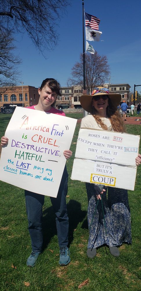 Photo of Evie and eepicheep with our protest signs. You can see the flags on the pole behind us blowing in the wind.

Evie's sign says, "America First" is CRUEL, DESTRUCTIVE, HATEFUL - - the LAST thing any _decent_ humans are about.

My sign is that poem I wrote on fb a while ago:

ROSES are RED,
Except when they're BLUE 
They call it "efficiency"
But it's truly a COUP 