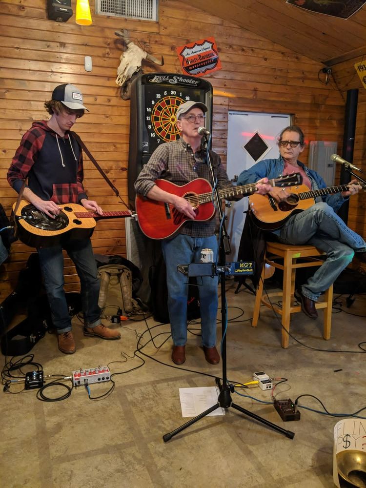 Three guys playing acoustic guitars. Guy at left standing, playing wood resophonic with a slide; middle also standing, singing into a mic; and at right, sitting with one leg crossed as a guitar rest and absolutely exuding chill. 

(Photo ca. 2020 by Bird, who is on here but I can't remember what he goes by.) 