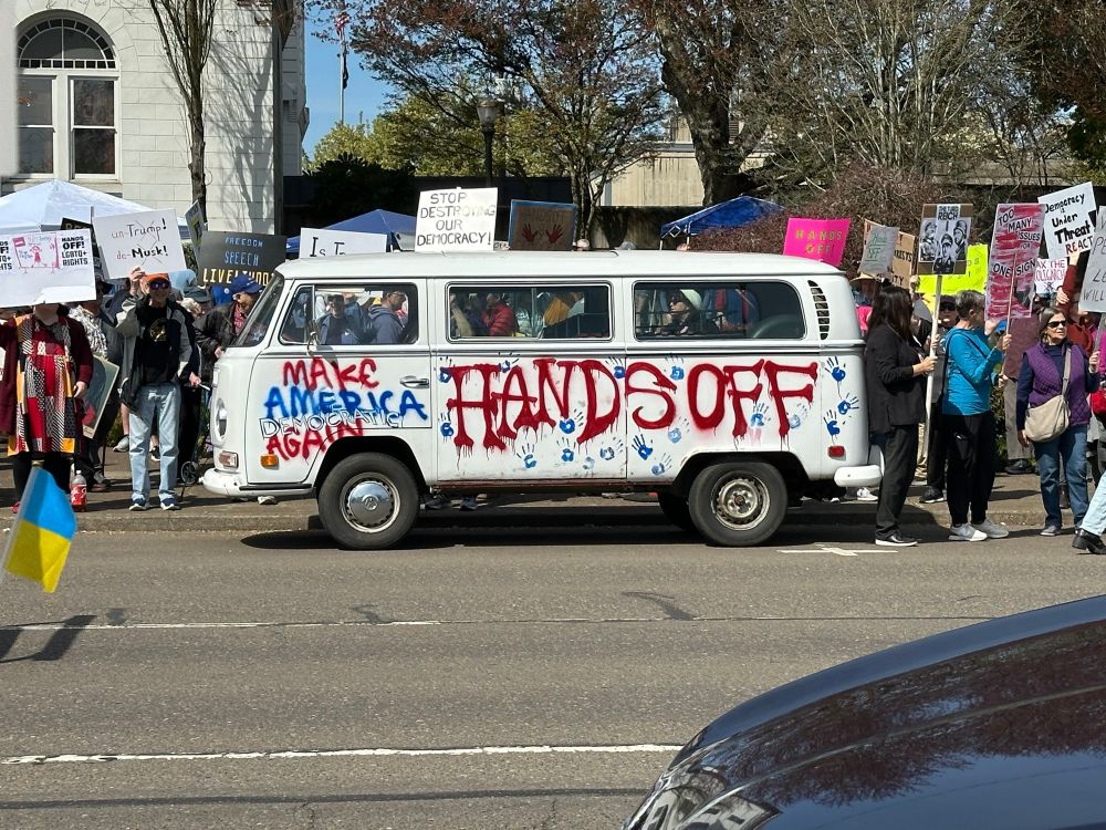 An image posted by someone here on bluesky showing a white VW bus with blue handprints "HANDS OFF" in red paint, and "Make America democratic again" on the driver's side, participating in the #handsoff demonstrations on Saturday, 5 April, 2025. Protesters holding signs are also part of the photo. Can you please let me know who posted so I can give photo credit? Thanks. 🤞😭