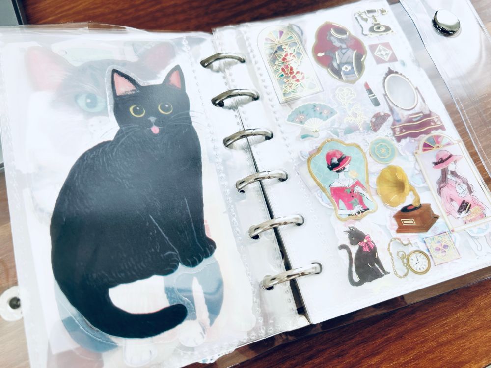 シール帳を開いたところ。左ページに大きな黒猫のステッカー、右ページにレトロ風の女の子や蓄音機、化粧台、花などのシリーズシール。