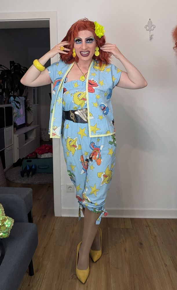 Ich posiere in dem Muppetfiguren Jumpsuit. Die Grundfarbe ist hellblau, verteilt sind die Muppetfiguren, die aus Sternen Fenstern winken. Ich trage gelbe Pumps und gelben Schmuck.