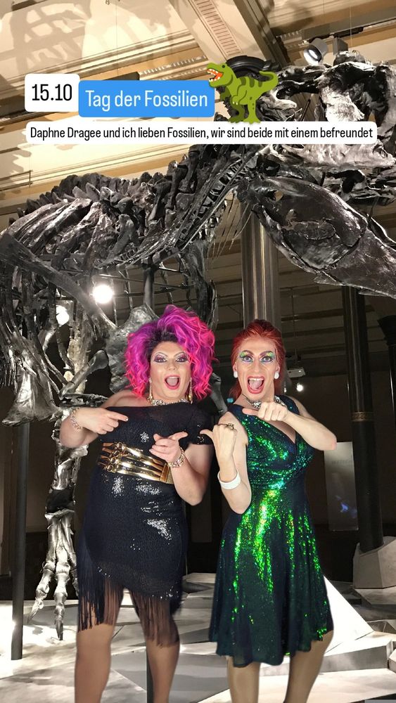 Daphne und ich stehen in funkelnden Kleidern in einem Museum vor einem großen Dinosaurierskelett und zeigen begeistert aufeinander.