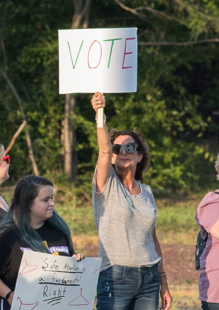Curly haired brunette woman wearing sunglasses, a grey T-shirt and jeans holding a sign at full arm extension that says, "VOTE"