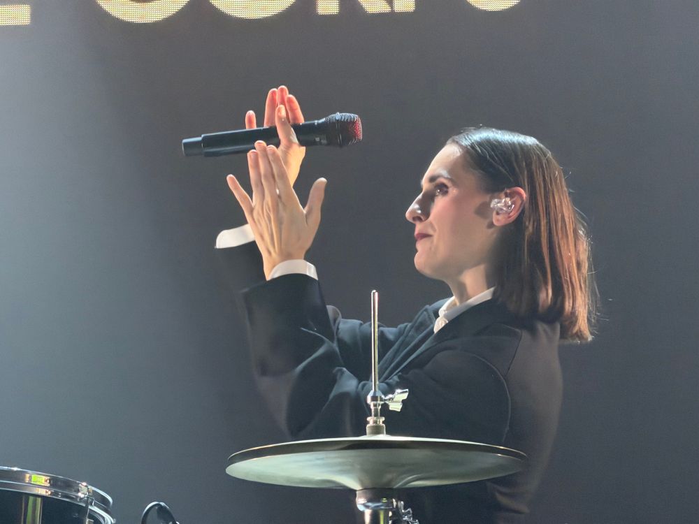 En gros plan sur une scène sombre, une personne est montrée de profil droit, concentrée, dans un moment précis de sa performance. Elle porte une veste noire structurée avec une chemise blanche visible au niveau du col et des poignets. Ses cheveux bruns, lisses et coupés au carré, sont soigneusement peignés derrière l’oreille.

Ses deux mains sont levées devant son visage, encadrant un micro maintenu à l’horizontale, comme si elle l’observait ou le mettait en scène avec précision. On distingue un écouteur transparent dans son oreille, soulignant le contexte de concert en direct.

Au premier plan, une cymbale de batterie apparaît dans la partie basse de l’image, accompagnée de sa tige métallique, ajoutant un élément instrumental à la composition. En arrière-plan, un écran sombre avec des lettres lumineuses partiellement visibles crée un contraste, renforçant l’esthétique professionnelle et immersive de la scène.

L’ensemble dégage une impression de maîtrise, de silence suspendu et d’intensité maîtrisée, comme juste avant ou après un moment musical clé.
