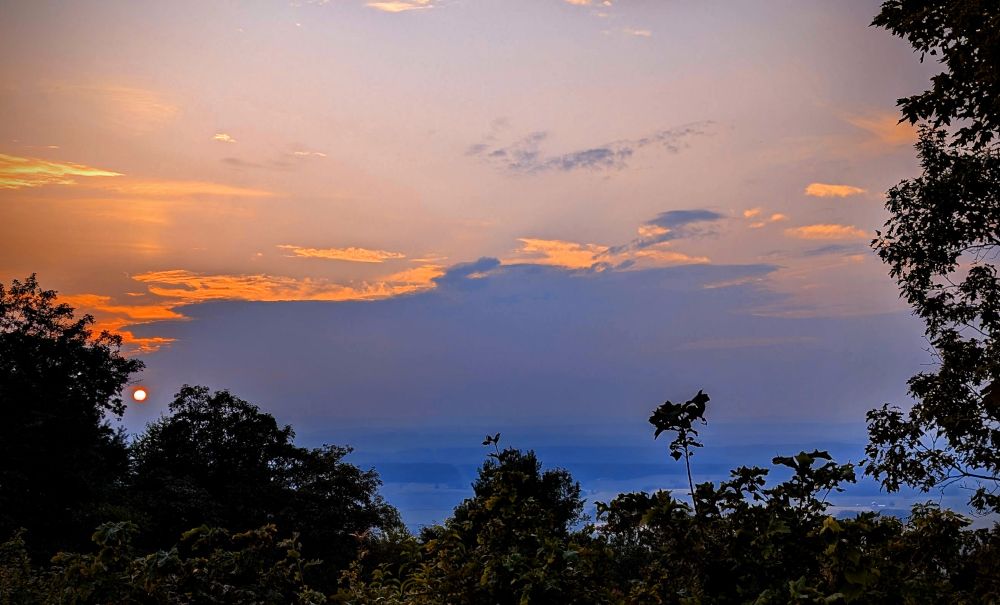 A sunset looking over a valley. Dark trees in the foreground. There are a series of blue hazy ridges disappearing into the distance.