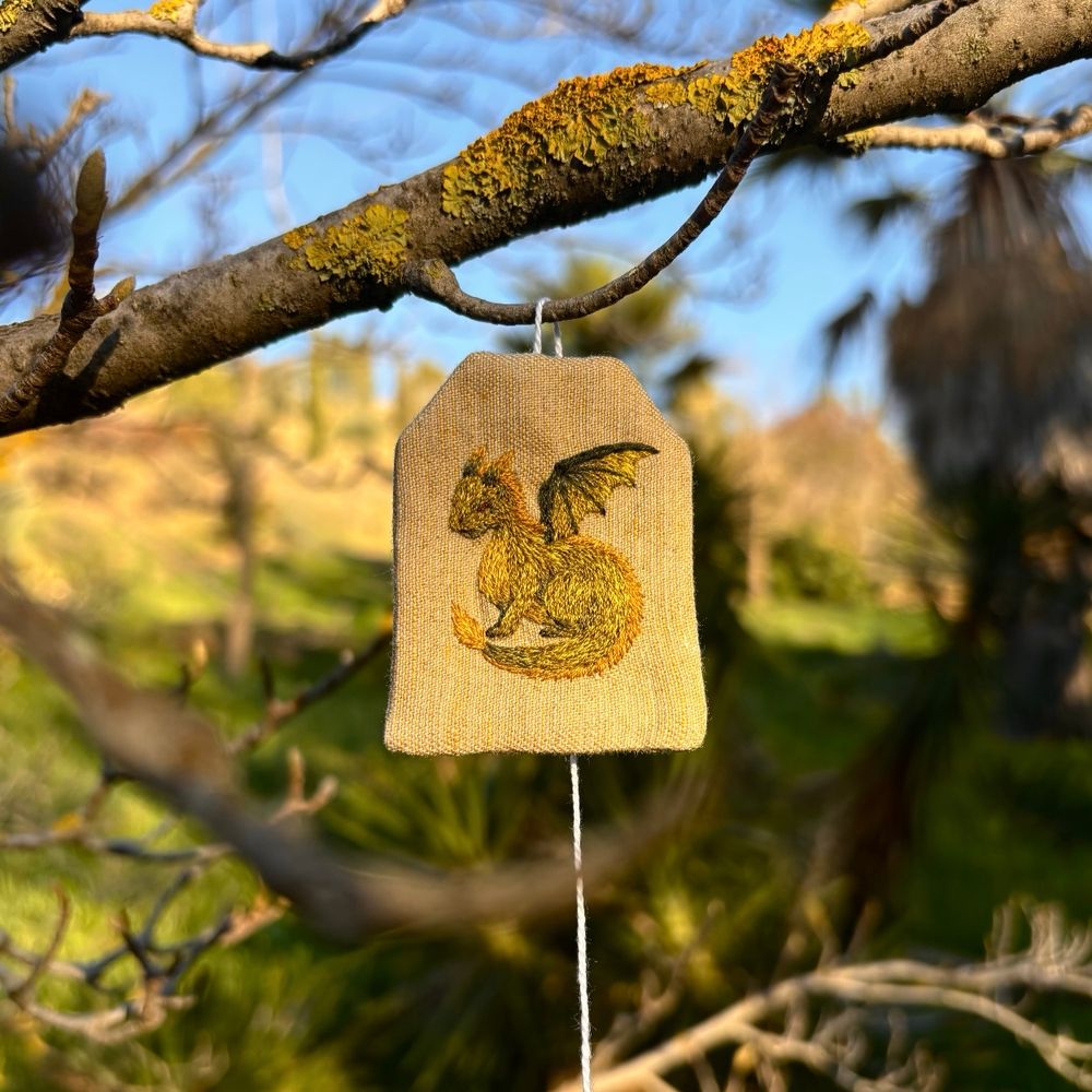 Fabric bookmark in the shape of a tea bag, with a green dragon embroidered on it. The bookmark is hanging from a tree branch.