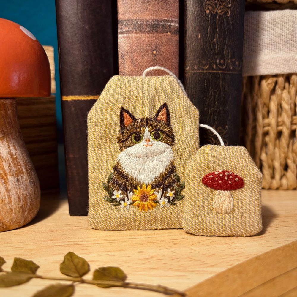 Bookmark shaped as a tea bag, with a cat embroidered on it. There is a small tag, just like in real tea bags, with a red mushroom embroidered on it.