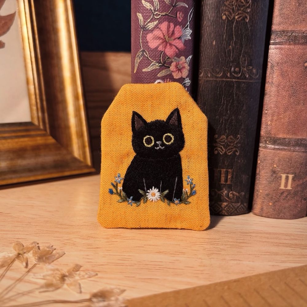 Bookmark made out of yellowish fabric, shaped as a tea bag. There is a black cat embroidered on it.