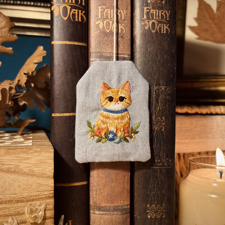 Blue fabric bookmark in the shape of a tea bag, with an orange cat embroidered on it. There are floral decorations as well.
