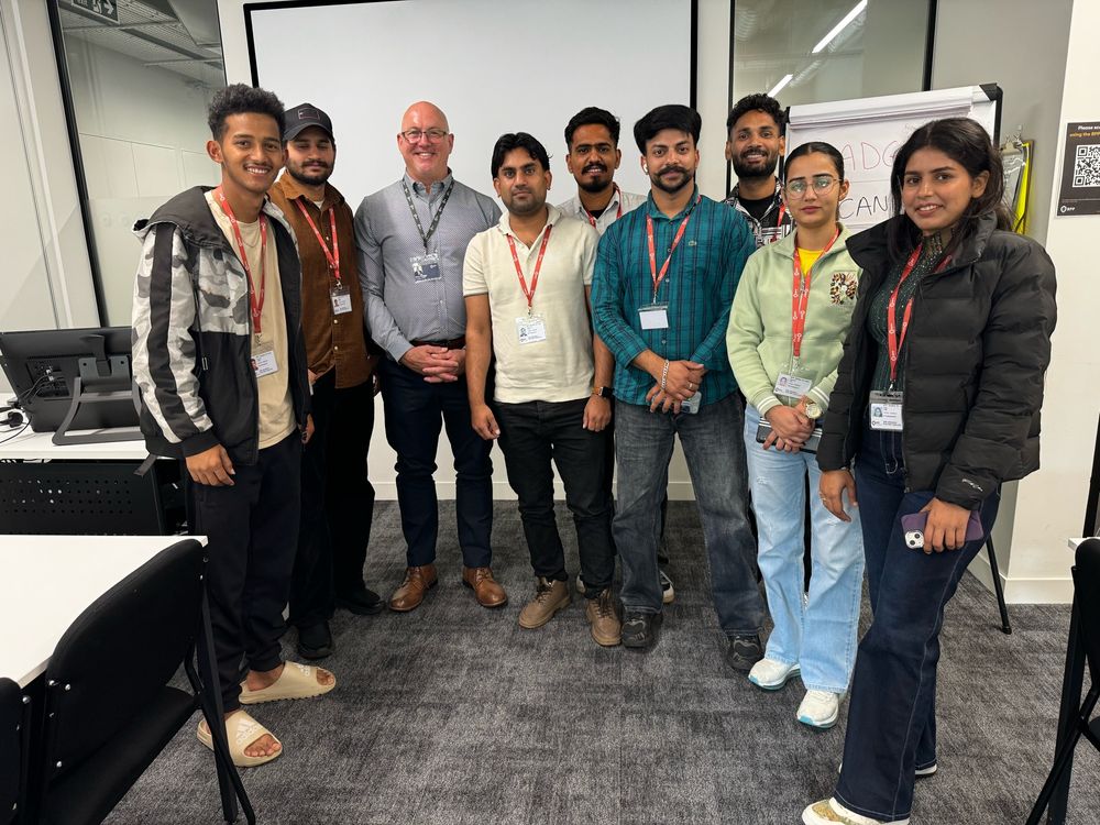 A mixture of international students, with their UK lecturer, having photo taken in front of a projector screen. Everyone is smiling.