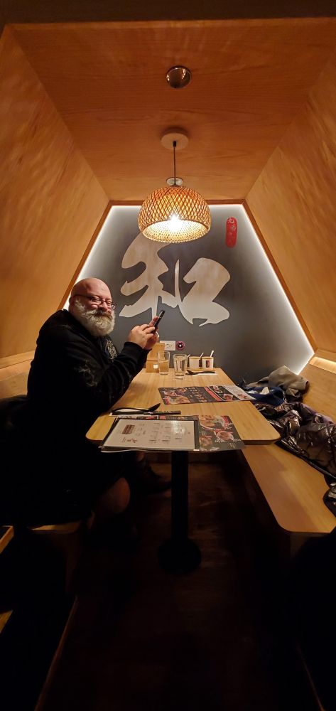 A hexagonal dining nook with a bald, great bearded man sitting on the left side and holding a smartphone. Menus are set on the edge of the table and winter coats are on the right-side bench.