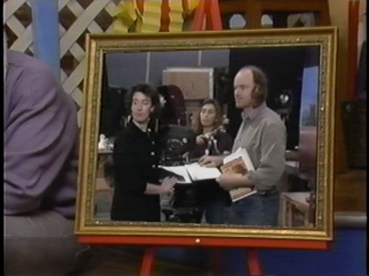 A still from "The Big A" episode 5 ("Teamwork"): Don Arioli sits (with only the profile of his left shoulder and thigh in-frame ) to the left of a picture fame. Superimposed within the picture frame is footage of Phil Tippet, with two unnamed female(?) crew workers, discussing their work on the set of a stop-motion film that the crew is working on.
