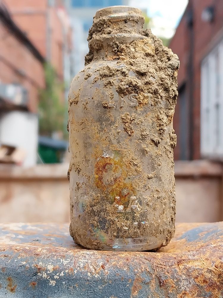A tiny (thumb size) fully intact glass bottle with dirt obscuring the makers Mark. The bottle rests on the edge of a dumpster at one end of an alley.