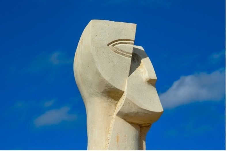 Skulptur eines Menschenkopfes aus hellem Stein gehauen. Der Kopf ist zweigeteilt und leicht verschoben: Hinterkopf und Auge und versetzt dazu Kinn und Nase. Im Hintergrund blauer Himmel. Skulpturenpark Zypern.