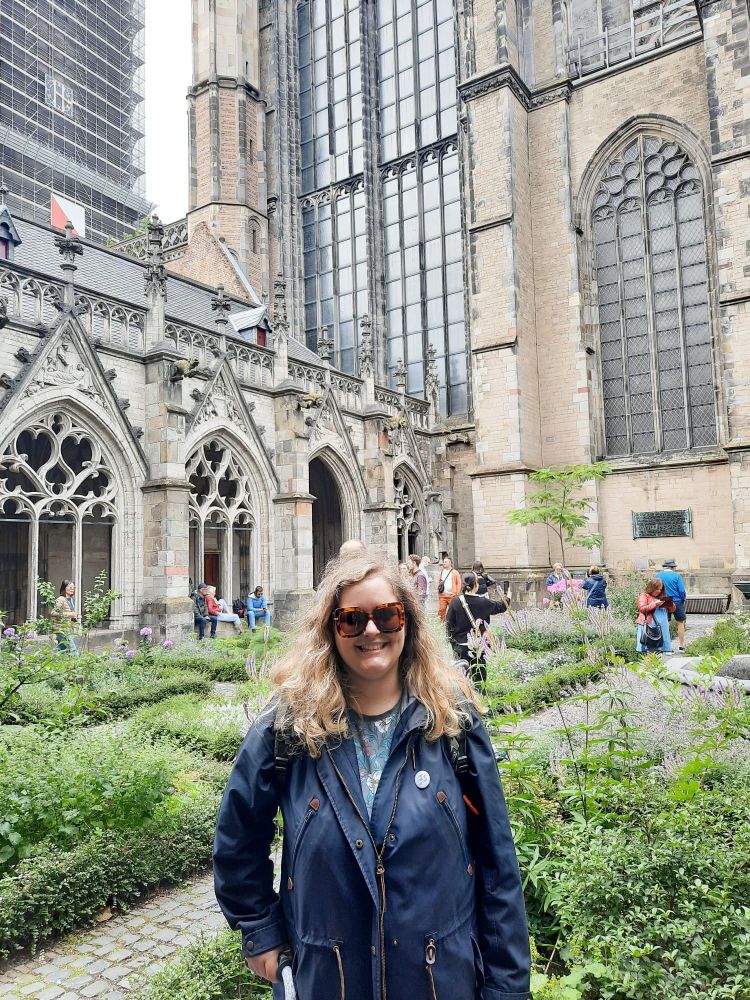 Photo. A white woman with wavy blonde hair and big sunglasses smiles. She is standing in a cloister of Utrecht Cathedral - garden plants and Gothic architecture are visible behind her. She is standing with a cane and wearing a blue jacket.