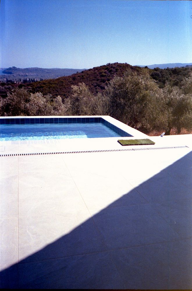 A colour photo with rectangular swimming pool under a clear blue sky. In the foreground is a light-colored tiled deck, partially enveloped by a large triangular shadow. Beyond the pool, there is a range of rolling hills covered with dense foliage 
