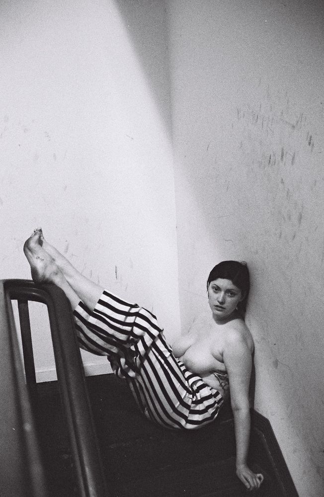 Monty is sitting in the stairwell, topless, striped pants, her legs up  resting on the handrail, bare feet. Black and white analogue portrait.
