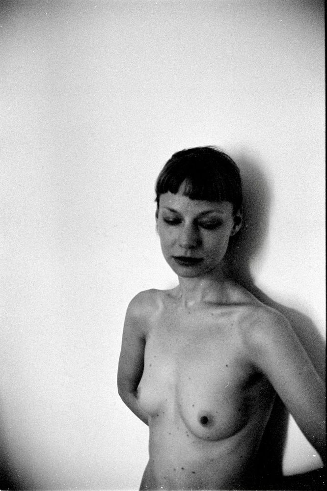 Black and white analogue photo of a dancer leaning with her back on a wall. She is topless, her body posture and her gaze suggest a contemplative state.  Perhaps in her mind she is playing the philosophical lyrics and slightly hypnotic rhythm of a favourite song, and some recent events in her life