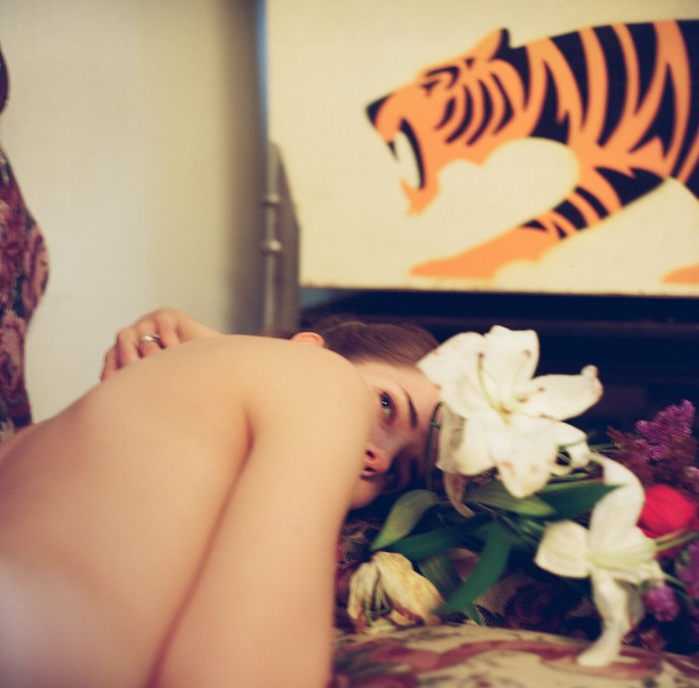 "The image features a close-up view of a woman lying on her side, with an arrangement of vibrant flowers including white lilies next to her face. The face is partially visible, with eyes looking beyond the camera. Behind her is a depiction of a stylized orange and black tiger, positioned with its mouth open as if roaring. The background includes a section of a patterned, multicolored fabric upholstery."


