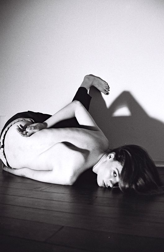 A black and white analogue image of a woman laying on the floor on her stomach, her body twisted to form a sculpture. We can see her face,  her lips. One eye is covered by her long hair. One hand rests on her nude back. One slender foot with painted toenails is pushing on the wall behind her, where the shadow reflects the composition.