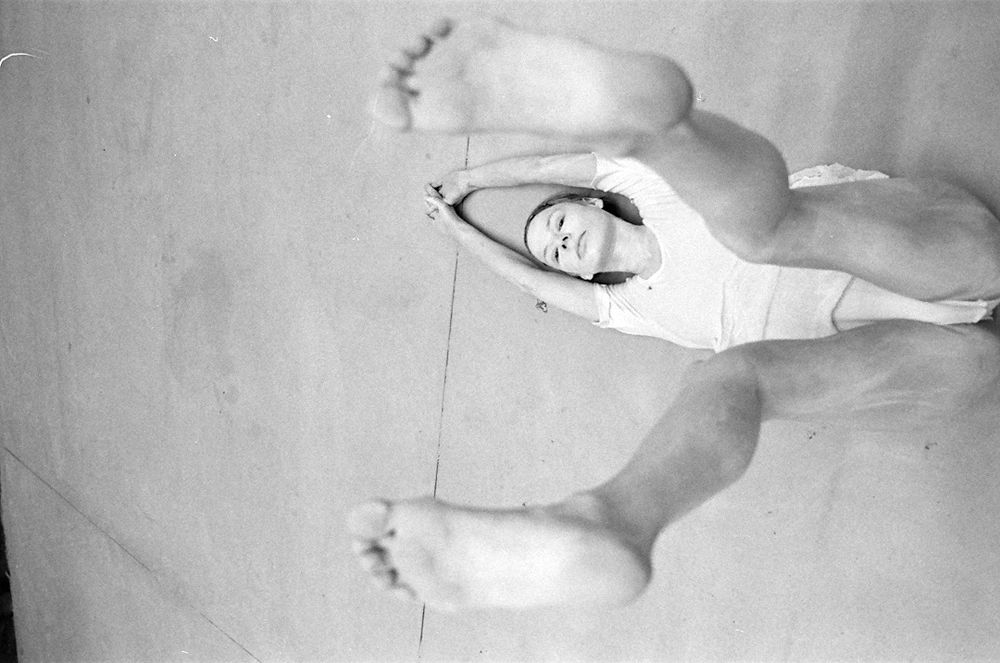 Black and white photo of a dancer lying on  her back with arms extended above the head and legs raised towards the camera, her bare feet in the foreground. There are traces of white paint on the face and body. The backdrop is a simple, featureless floor with a faint diagonal line visible. 
©RayMax