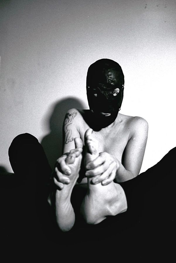 Black and white analogue photo of a seated woman wearing a gimp mask, topless, hands holding her bare feet up in front of the camera