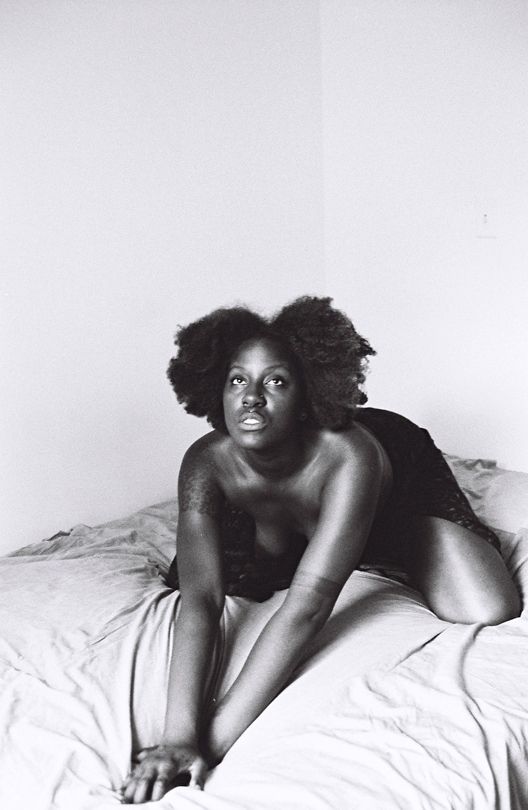 "Black and white photo of a Black woman with natural, voluminous hair, kneeling on a bed with both arms stretched forward and their fingers spread on the sheets. The lighting natural, the woman's gaze is directed slightly upwards"