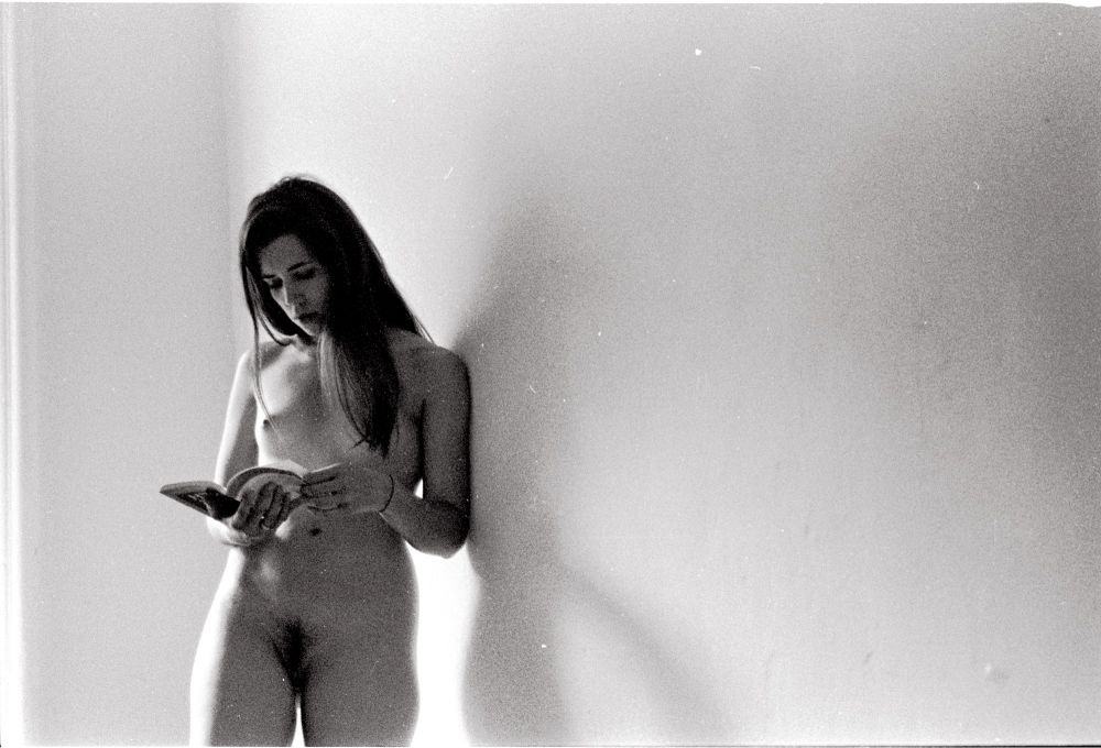 Melanie stands nude against a blank wall enjoying a poetry book. Black and white analogue photograph