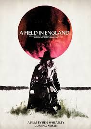 A Field in England film, a black and white figure in a cloak and hat walks across a field under a red sun.