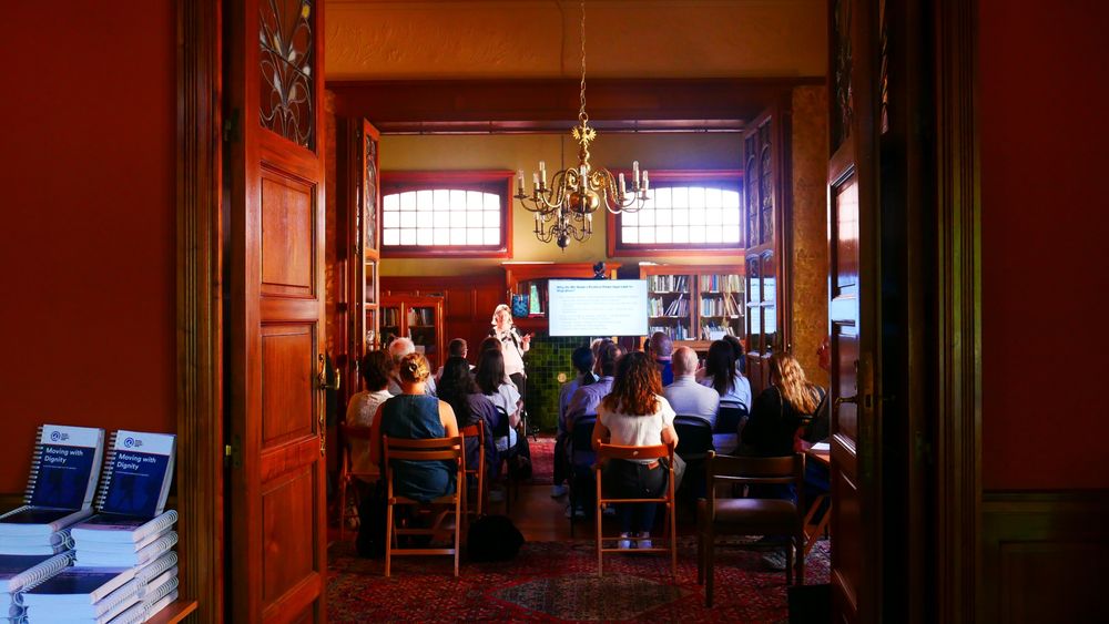 An interior of a Quaker House, Brussels. A group of people listens to a female speaker talking about a recently launched publication. 