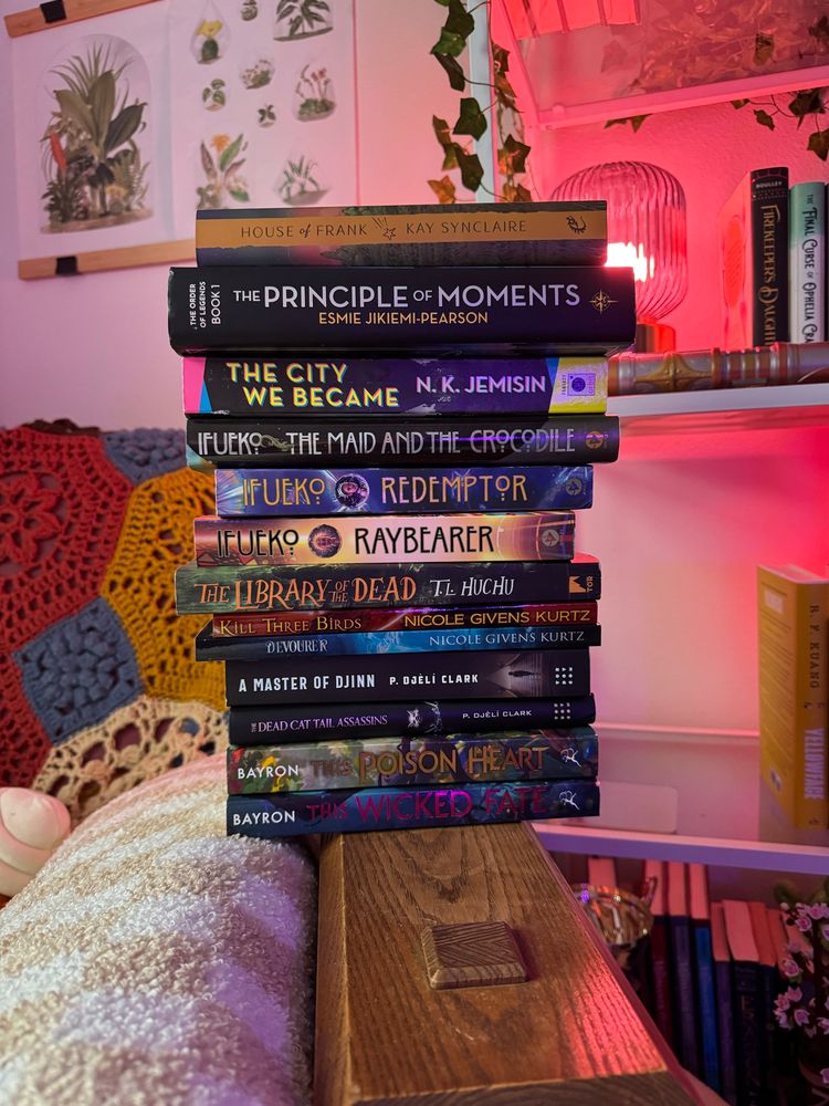 A stack of books sits on the arm of a chair. The books are: House of Frank by Kay Synclaire, The Principle of Moments by Esmie Jikiemi-Pearson, The City We Became by N. K. Jemisin, The Maid and the Crocodile by Jordan Ifueko, Redemptor by Jordan Ifueko, Raybearer by Jordan Ifueko, The Library of the Dead by T. L. Huchu, Kill Three Birds by Nicole Givens Kurtz, Devourer by Nicole Givens Kurtz, Master of Djinn by P. Djeli Clark, The Dead Cat Tail Assassins by P. Djeli Clark, This Poison Heart by Kalynn Bayron, This Wicked Fate by Kalynn Bayron.