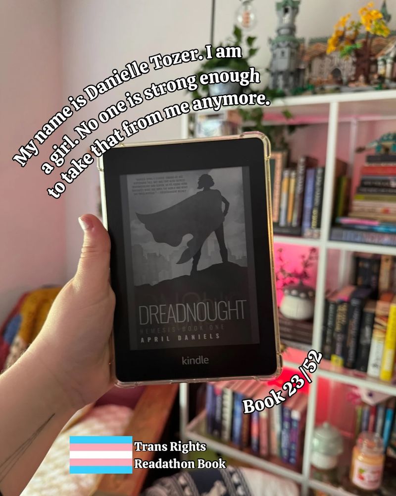 a hand holds a kindle in front of a bookshelf. The kindle displays a black and white cover of Dreadnought by April Daniels. Text on the image reads “My name is Danielle Tozier. I am a girl. No one is strong enough to take that from me anymore,” “Book 23/52,” and “Trans Rights Readathon Book.”