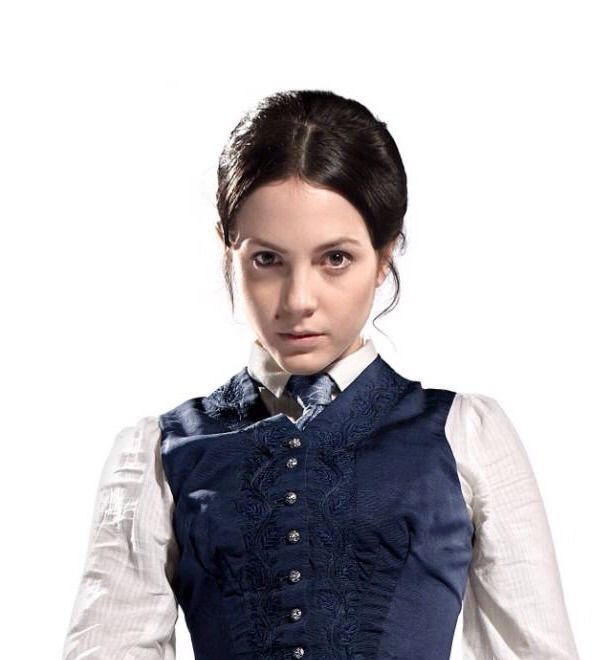 Uma foto da personagem Jenny Flint da série Doctor Who. Jenny é uma mulher branca, de cabelos castanhos escuros amarrados, e está usando blusa branca de manga longa por baixo de um vestido azul marinho de botões.