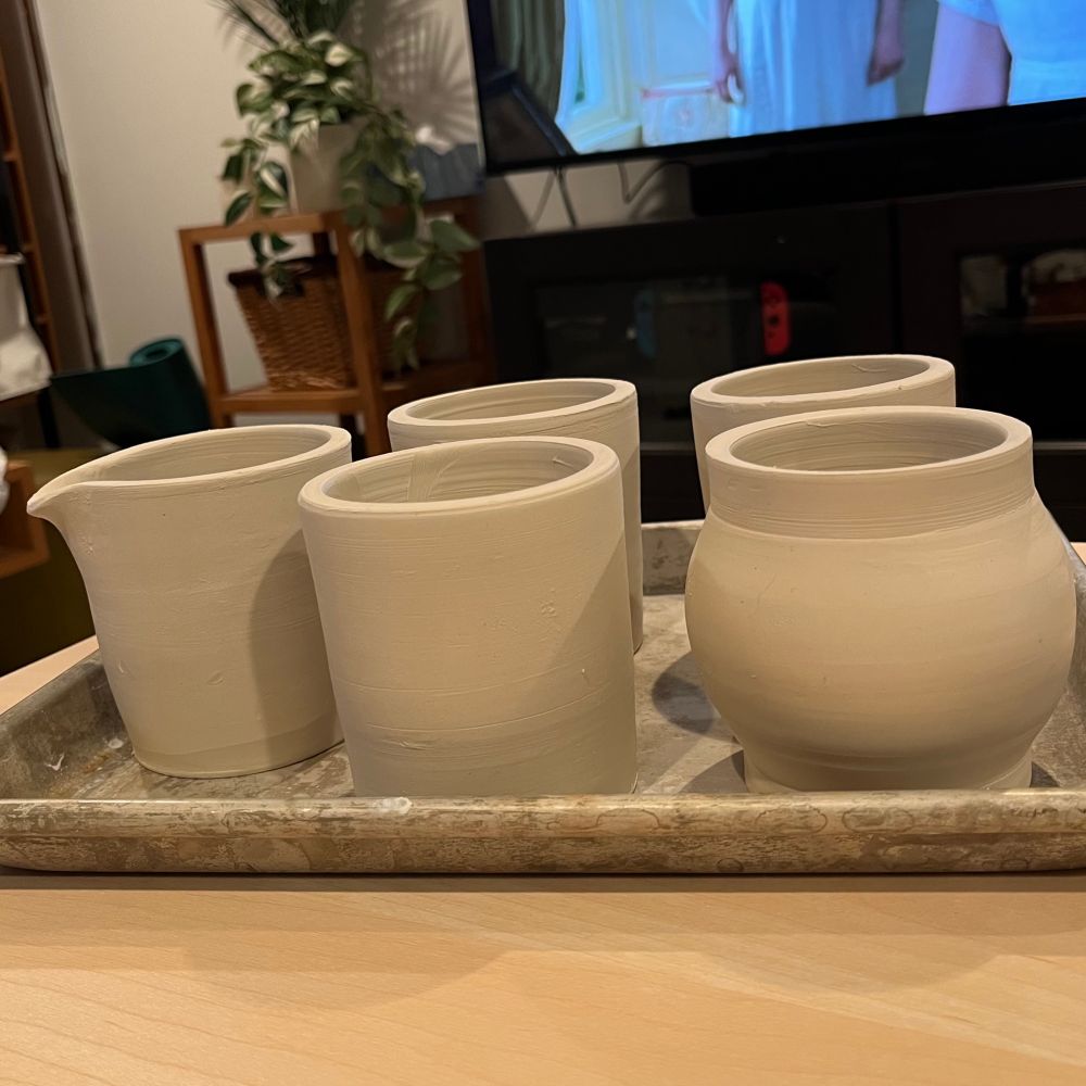 5 hand-thrown porcelain vessels on a tray. Their surfaces are plain, ready for carving. A TV is on in the background. 