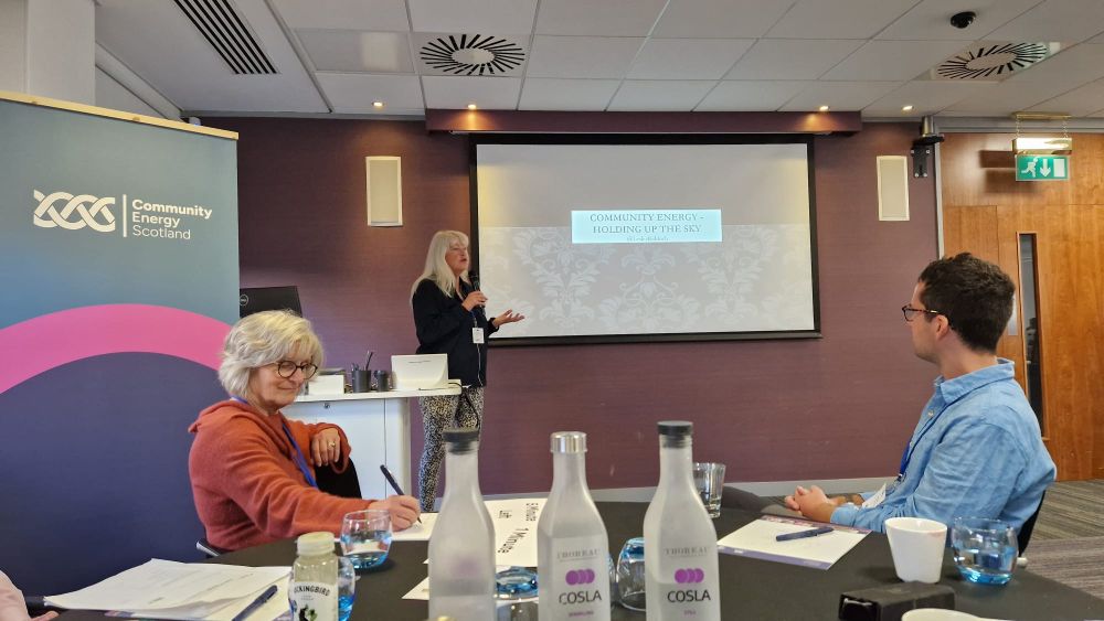 Two people sit at a round table. Water bottles in the foreground. A branded banner on the left. A woman with long white hair is speaking, in front of a big presentation screen.