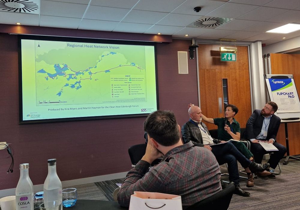 Three people are sitting on chairs on the right side of the shot.  A presentation screen is to the left, and a woman in a chair is gesturing back towards it. A man sits on a chair facing away from the camera. The wall behind the screen is aubergine coloured. The screen has a map on it.
