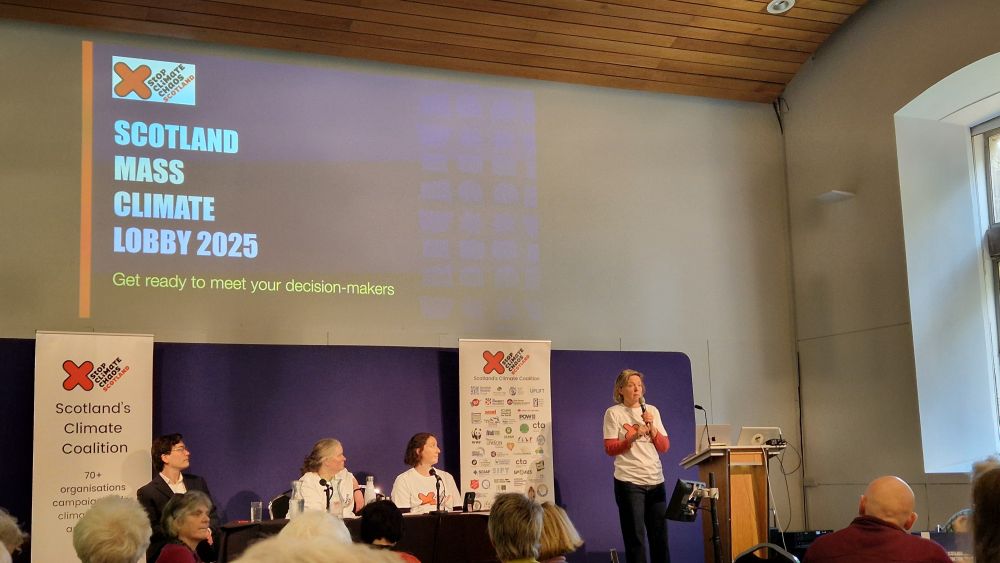 4 people are on a stage. One woman is standing and holding a mic, behind a podium. Projected on the wall it says "Scotland Mass Climate Lobby".