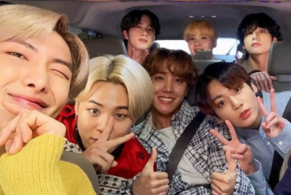 The 7 members of BTS—RM, Jin, Suga, J-hope, Jimin, V, and Jungkook—riding in a car. RM in the front; Jimin and Jungkook holding peace signs while J-Hope give 2 thumbs up are in the middle row; and Jin, Suga, and V are in the back row. Image take from their Carpool Karaoke segment.