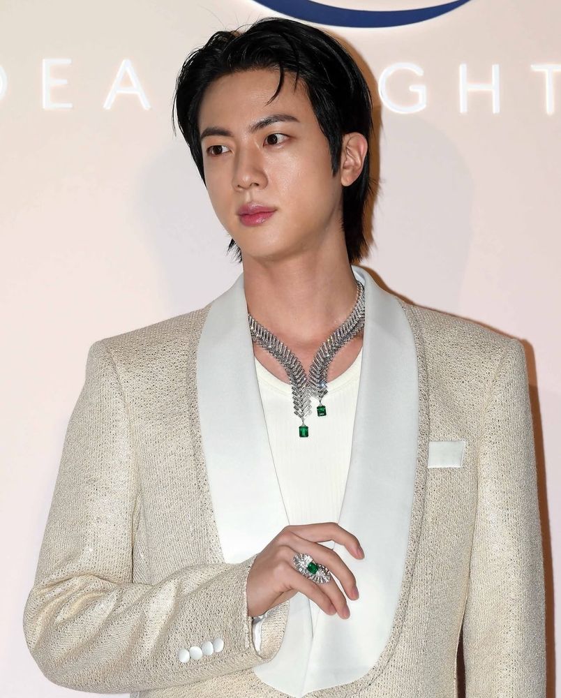 Kim Seokjin of BTS at a Maison FRED event wearing a sequined champagne-colored tuxedo jacket with satin lapels over a cream colored top. In his neck is a diamond and emerald necklace which matched the ring on his right hand ring finger.